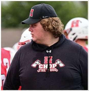 People wearing red sports jerseys stand near a person in a black hoodie with a graphic that reads “CHOP THE WOOD" and features two axes. Description generated by AI