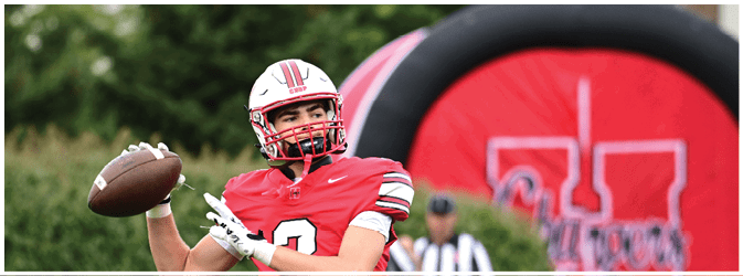 A person wearing a red football uniform and helmet is preparing to throw a football on a field with a large red and black inflatable structure in the background. Description generated by AI