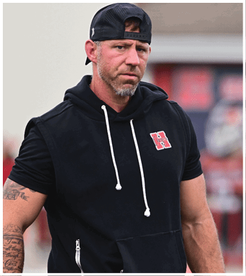 A person wearing a sleeveless black hoodie with a red “H" logo and a black backward cap is standing with a serious expression and visible tattoos on their arm. Description generated by AI