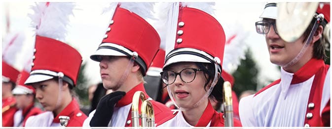 People wearing red marching band uniforms and tall red hats are standing close together, some holding brass instruments. Description generated by AI