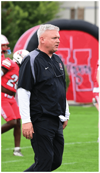 A person with short gray hair wearing a black sports jacket and pants stands on a grassy field with players in red and white football uniforms in the background. Description generated by AI