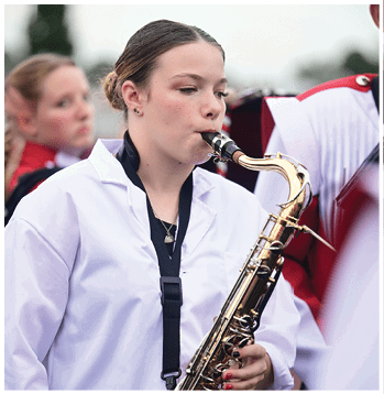 A person wearing a white uniform is playing a saxophone outdoors with other people in similar attire visible in the background. Description generated by AI