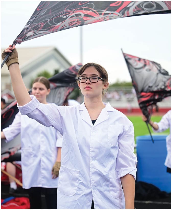 A person wearing glasses stands outdoors under a patterned umbrella with another person partially visible in the background. Description generated by AI
