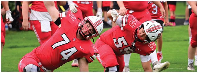 Two football players in red uniforms and helmets are stretching on a field while other players and people stand in the background. Description generated by AI