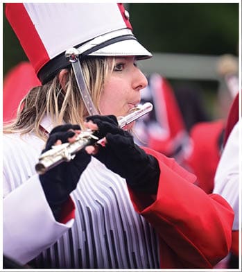 A person wearing a red and white marching band uniform and hat is playing a flute outdoors. Description generated by AI