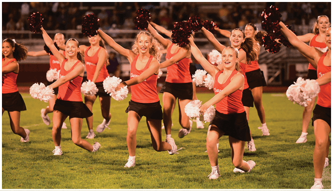A group of adults wearing red tops and black skirts perform a cheerleading routine on a grassy field while holding white and dark red pom-poms. Description generated by AI