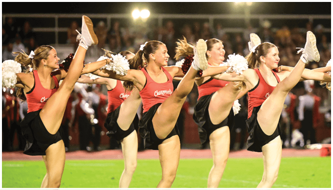 A group of adults wearing red tops and black skirts are performing a high kick routine while holding white pom-poms. Description generated by AI