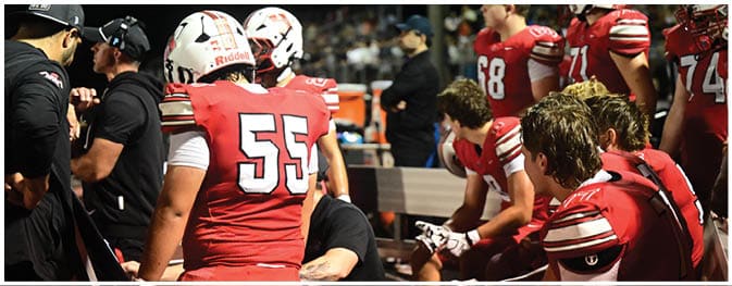 People wearing red and white football uniforms are gathered on the sidelines during a game, with some sitting on a bench and others standing. Description generated by AI