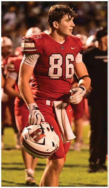 A person wearing a red football uniform with the number 86 is holding a white helmet while standing on a grassy field with other players in the background. Description generated by AI