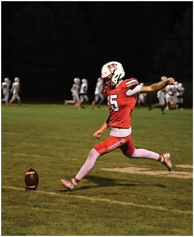 A person wearing a red sports uniform and white helmet is kicking a football on a grassy field at night with other players visible in the background. Description generated by AI