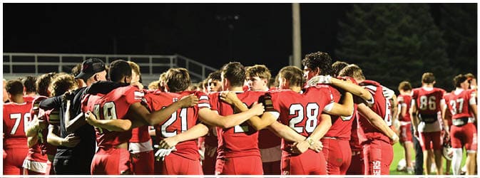 A group of people wearing red sports uniforms are standing in a close huddle on a field at night. Description generated by AI