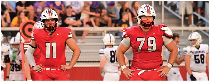Two people wearing red football uniforms and helmets stand on a field with spectators in the background. Description generated by AI