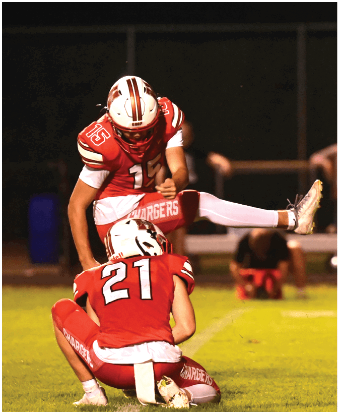 Two people in red football uniforms are on a field at night with one holding the ball on the ground while the other prepares to kick it. Description generated by AI