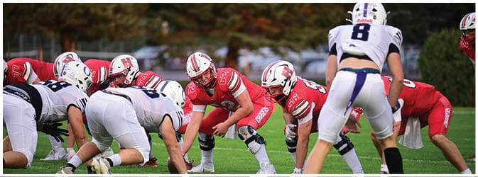 People in football uniforms are crouched on a grassy field preparing for a play during a game. Description generated by AI