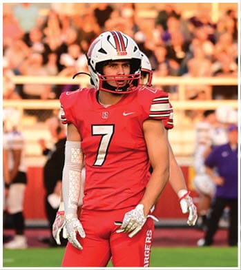 A person wearing a red football uniform with the number seven stands on a grassy field with other players and spectators in the background. Description generated by AI