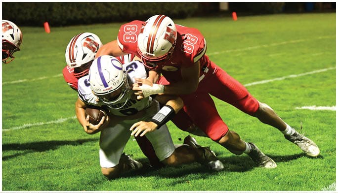 People in football uniforms are engaged in a tackle on a grassy field at night. Description generated by AI