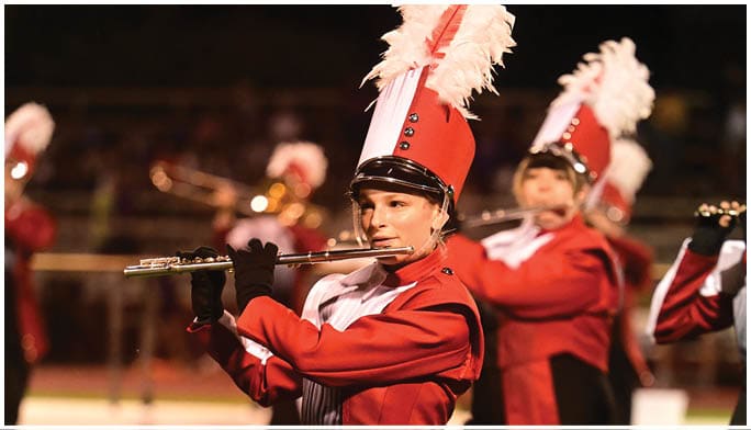 People wearing red and white marching band uniforms with tall hats adorned with white feathers are performing. Description generated by AI