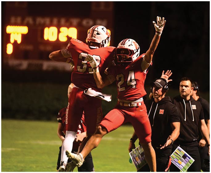 Two people in red football uniforms jump and celebrate on a field while three others in black clothing stand nearby holding clipboards. Description generated by AI