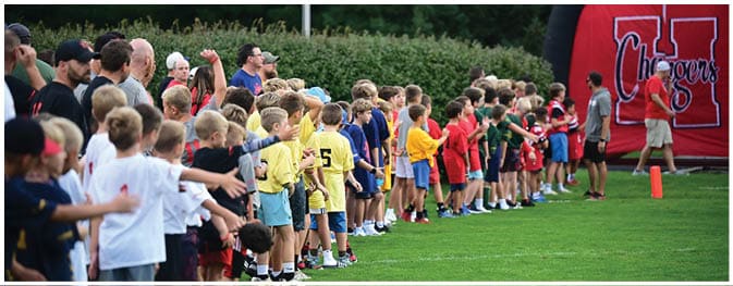 A large group of children wearing colorful sports jerseys stand in a line on a grassy field while adults watch nearby. Description generated by AI