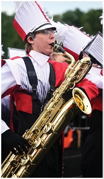 Person wearing a white and red marching band hat and glasses is playing a clarinet outdoors. Description generated by AI