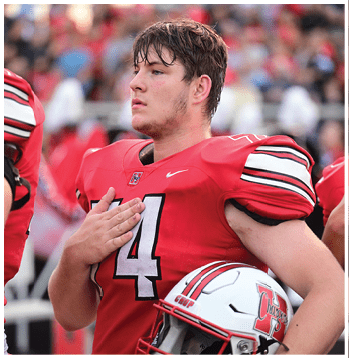 A person in a red football uniform with the number 14 holds a helmet and places a hand over their chest, standing in a stadium with a blurred crowd in the background. Description generated by AI