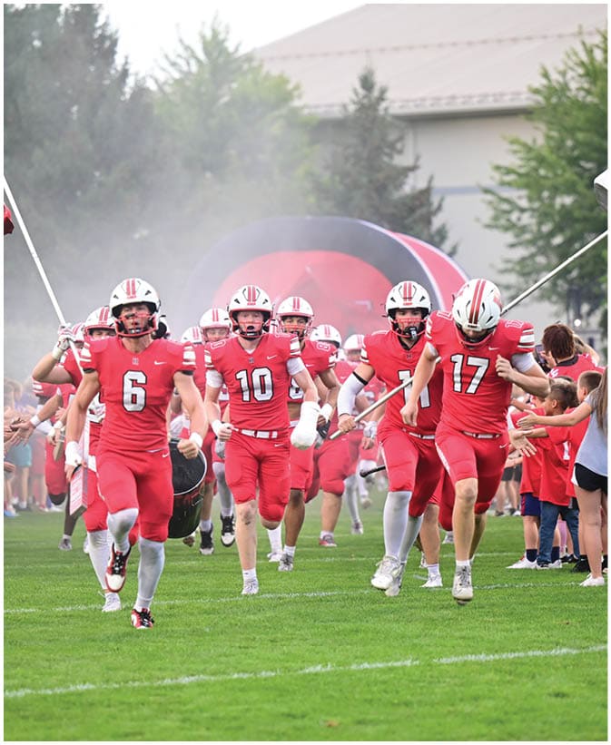 A group of people in red sports uniforms and helmets run onto a grassy field from a tunnel while holding flags and high-fiving a line of people standing on the sidelines. Description generated by AI