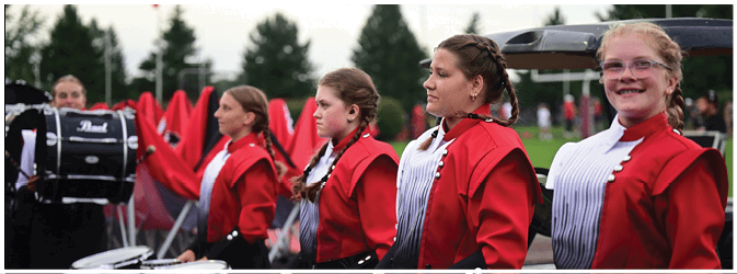 People wearing red and white marching band uniforms stand outdoors with musical instruments and flags. Description generated by AI
