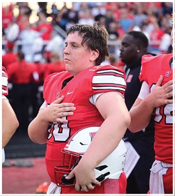 A person in a red football uniform holds a white helmet in one hand and places the other hand over their chest. Description generated by AI