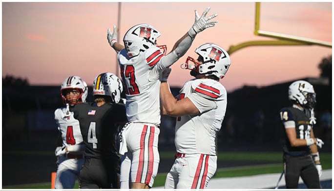 Two people in white football uniforms celebrate in an end zone while three people in black football uniforms stand nearby on a field at sunset. Description generated by AI