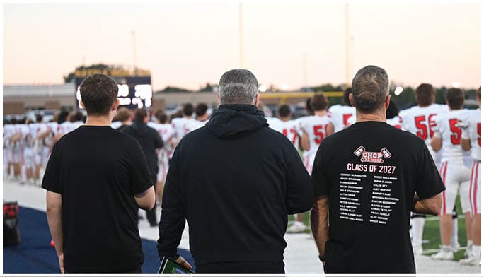 Three adults stand on the sidelines of a football field watching a line of players in white and red uniforms standing in formation during a game or practice. Description generated by AI