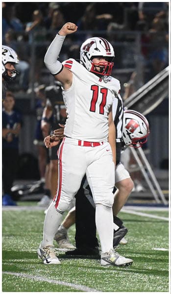 A person in a white football uniform with the number 11 raises a fist while standing on a football field surrounded by other players in dark uniforms. Description generated by AI