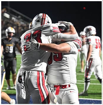 Two football players in white uniforms with red stripes embrace on a field while other players in black uniforms stand nearby. Description generated by AI