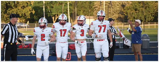 Four people in white football uniforms walk arm in arm on a field while two referees and another person in casual clothing stand nearby. Description generated by AI