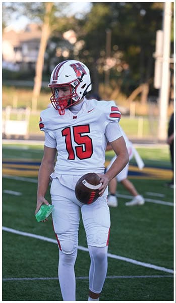A person wearing a white football uniform with the number 15 and a helmet is holding a football and a green item while standing on a football field. Description generated by AI