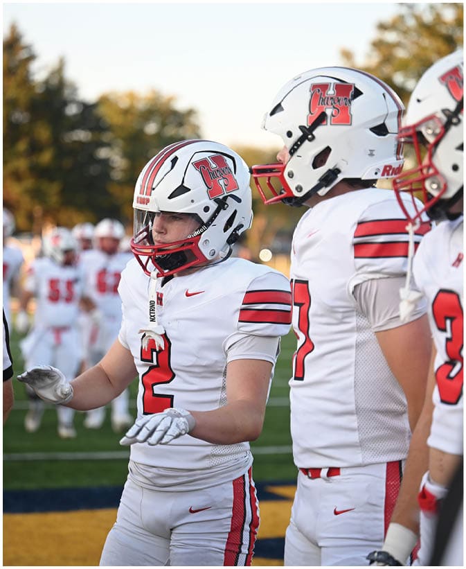 Two people wearing white football uniforms and helmets with red and black accents stand on a field with blurred players and trees in the background. Description generated by AI