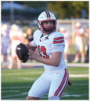 A person in a white football uniform with red and black stripes is holding a football and preparing to throw it on a green field with other players and spectators blurred in the background. Description generated by AI