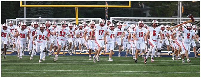 A large group of people wearing white sports uniforms with red numbers are running onto a football field from the sidelines. Description generated by AI