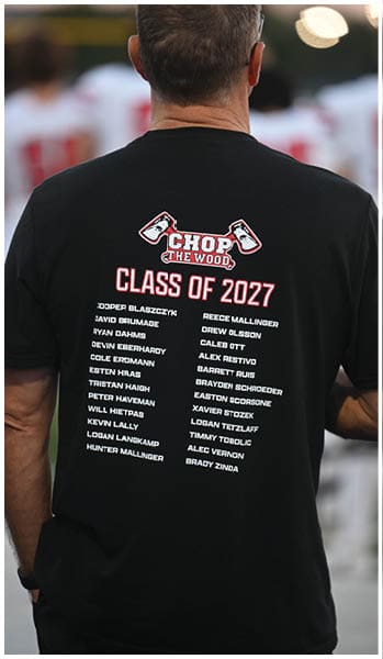 A person wearing a black t-shirt with a “Chop the Wood Class of 2027" design and a list of names on the back stands outdoors with blurred people in sports uniforms in the background. Description generated by AI