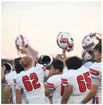 A group of people wearing white sports uniforms with red numbers are raising their helmets in the air on a field during sunset. Description generated by AI