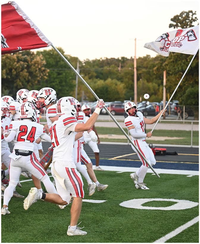 A group of people in white football uniforms with red and black stripes are running onto a field, some holding large flags. Description generated by AI