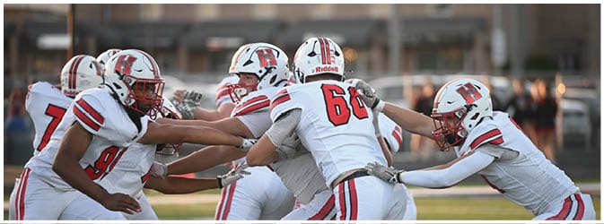People in football uniforms are engaged in a play on a field, with some reaching out to block or tackle. Description generated by AI