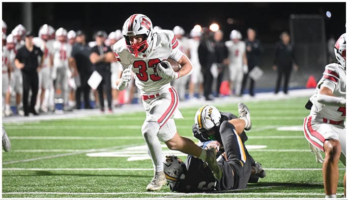 A person in a white and red football uniform is running with the ball while two people in dark uniforms are on the ground during a nighttime football game. Description generated by AI