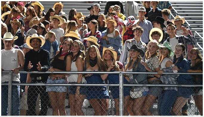 A group of people wearing cowboy hats and casual clothing are standing closely together behind a metal fence, smiling and posing for the camera at an outdoor event. Description generated by AI