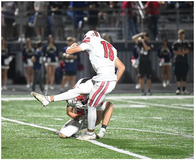 A person in a white sports uniform kicks a football while another person holds the ball on the ground during a nighttime game on a green field with spectators in the background. Description generated by AI
