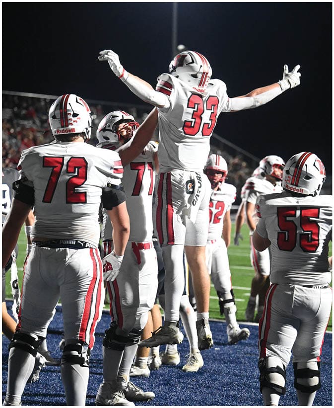 People wearing white football uniforms with red and black stripes celebrate on a field under bright stadium lights at night. Description generated by AI