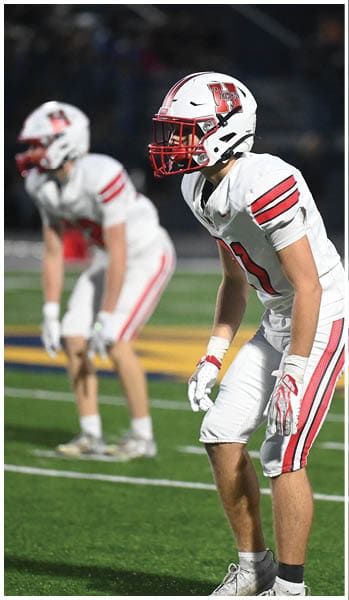Two people in white football uniforms with red stripes and helmets are positioned on a field, appearing ready for action during a game. Description generated by AI