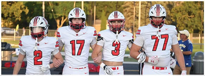 Four people in white football uniforms with red numbers walk arm in arm on a field with a blue track and a person in casual clothing in the background. Description generated by AI