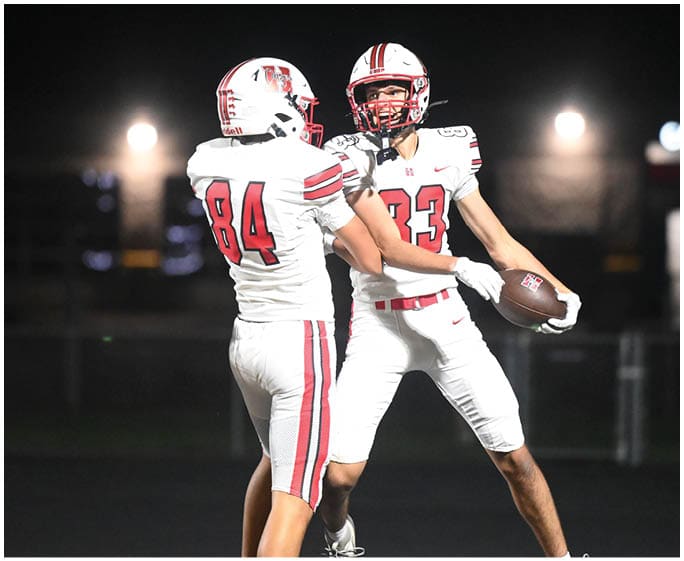 Two people in white football uniforms with red stripes celebrate on a field at night, one holding a football. Description generated by AI