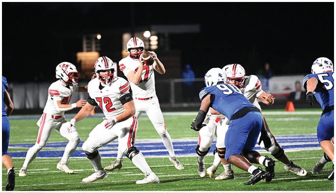 Players in white and blue football uniforms are actively engaged in a nighttime game on a field with artificial turf. Description generated by AI