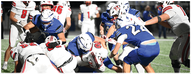 Several people wearing helmets and sports uniforms are engaged in a close physical struggle on a football field during a game. Description generated by AI